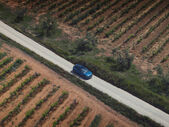Carro azul viaja por estrada ladeada por campos agrícolas.
