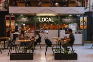 Pessoas almoçam no restaurante 'Local', com decoração de plantas e iluminação neon