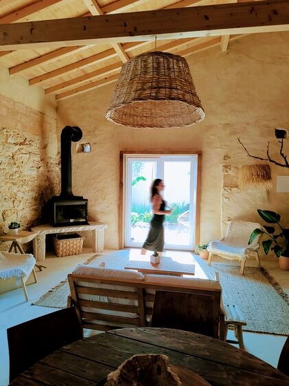 Sala rústica com lareira, cadeiras de madeira e uma mulher perto da janela