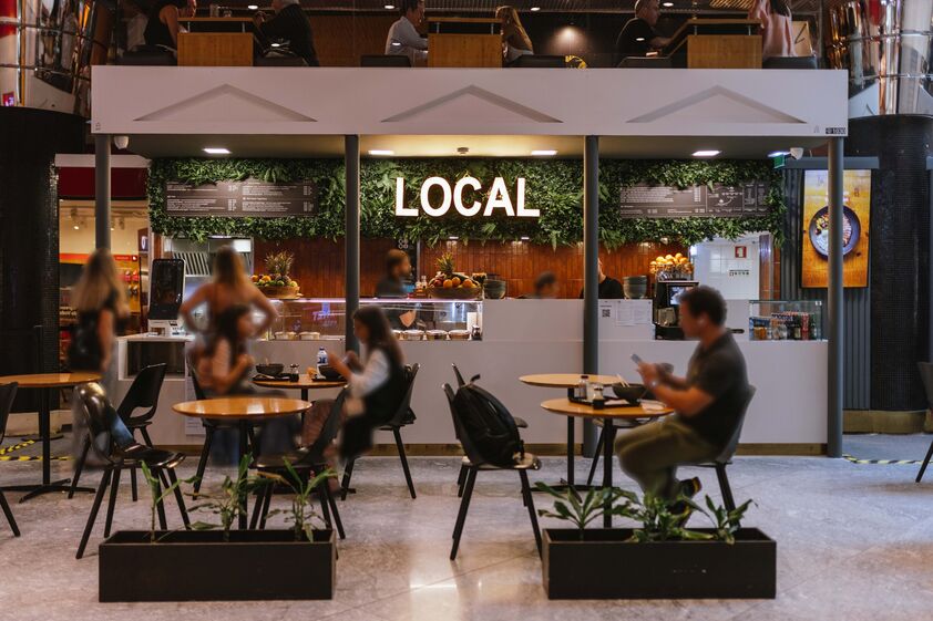 Pessoas almoçam no restaurante 'Local', com decoração de plantas e iluminação neon