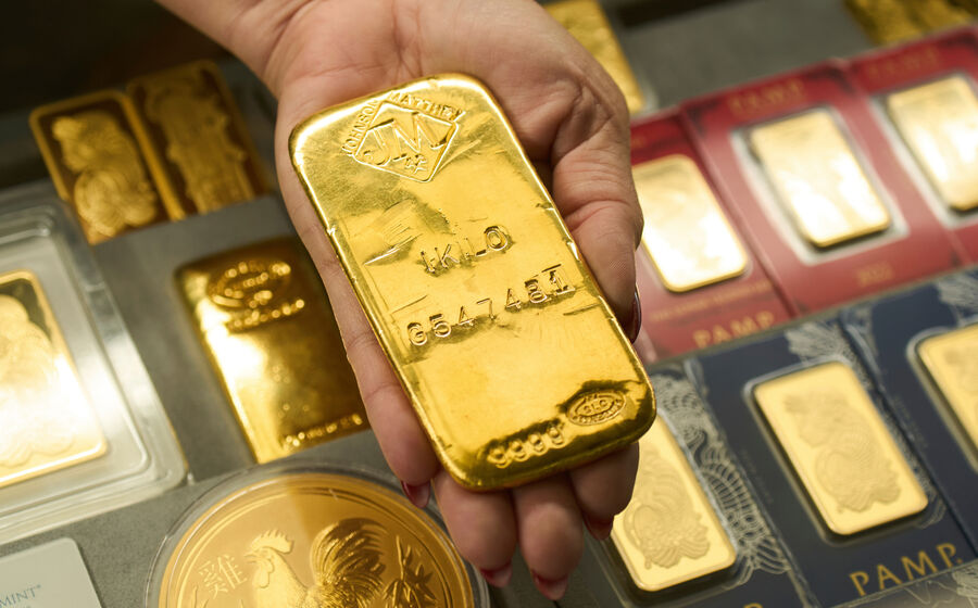 Barra de ouro de 1 kg na mão, com outras barras e moedas de ouro visíveis