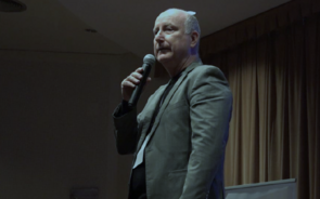 Homem discursa ao microfone num palco com cortinas