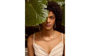 Mulher posa com colar e brincos dourados perto de uma grande folha verde